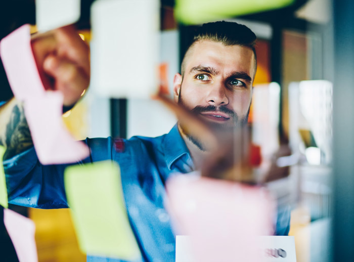 A focused young man in a blue shirt is analyzing colorful sticky notes on a glass board in an office environment, engaged in a brainstorming session or planning process.