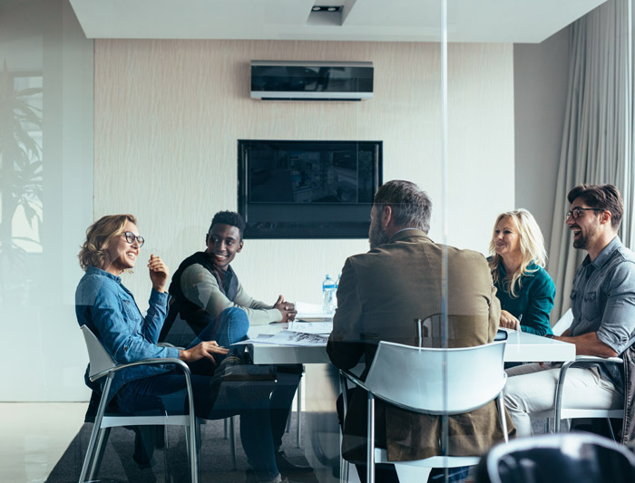 A group of diverse professionals engaged in a meeting around a conference table, with one person laughing and others smiling, in a modern office setting.