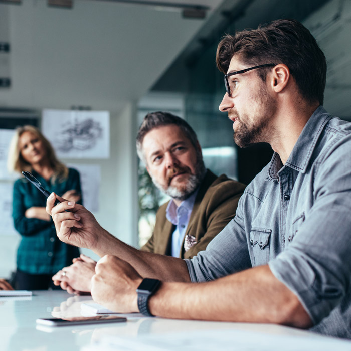 Three professionals engaged in a business discussion at a conference table, with one man gesturing while holding a pen and looking at another man, who is attentively listening. A woman stands in the background, appearing interested in the conversation.