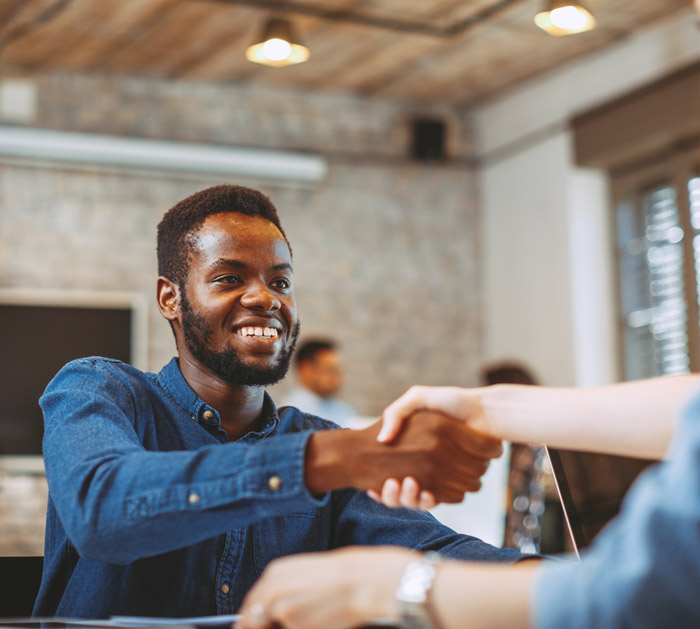 A smiling man in a blue shirt shakes hands with another person in a business setting, symbolizing collaboration and partnership.