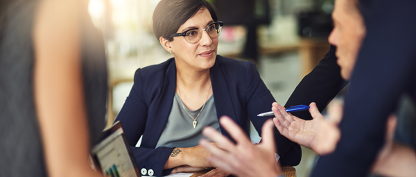 A woman wearing glasses engages in a discussion during a business meeting, while two men gesture animatedly around her. The setting appears professional and collaborative, emphasizing team dynamics.