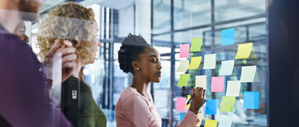 Two people observe a woman writing on colorful sticky notes attached to a glass wall in a modern office environment. The focus is on collaboration and brainstorming with post-it notes.