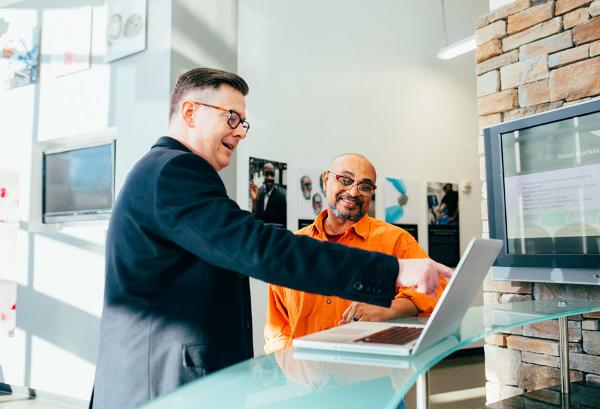 Two men engaged in a discussion at a modern office reception, with one man pointing at a laptop screen while the other smiles, highlighting professionalism and collaboration in a bright environment.