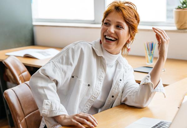 Young woman with red hair laughing while sitting at a desk, wearing a white shirt, in a bright workspace with a laptop and stationery.